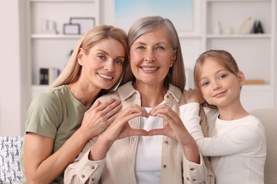 Photo of Three generations. Happy grandmother showing heart gesture with her daughter and granddaughter at home