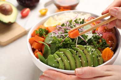 Photo of Woman eating delicious vegan bowl with avocados, carrots and tomatoes at light table, closeup