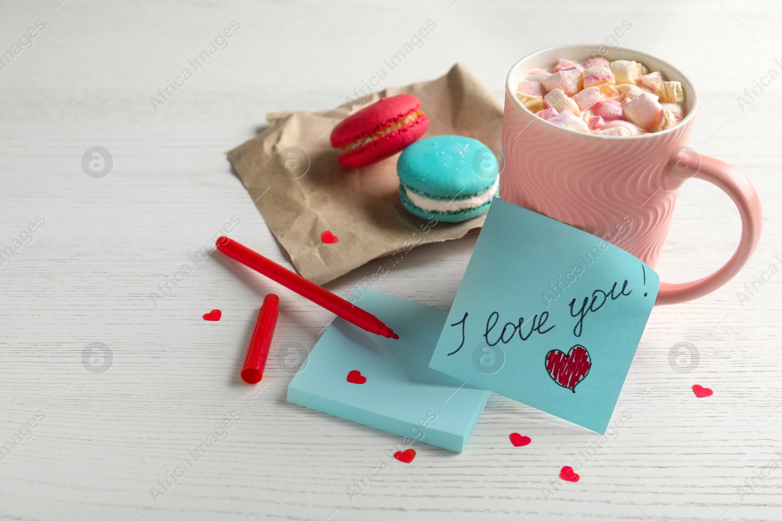 Photo of Note with phrase I Love You near pink cup of hot drink and macarons on white wooden table