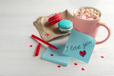 Photo of Note with phrase I Love You near pink cup of hot drink and macarons on white wooden table