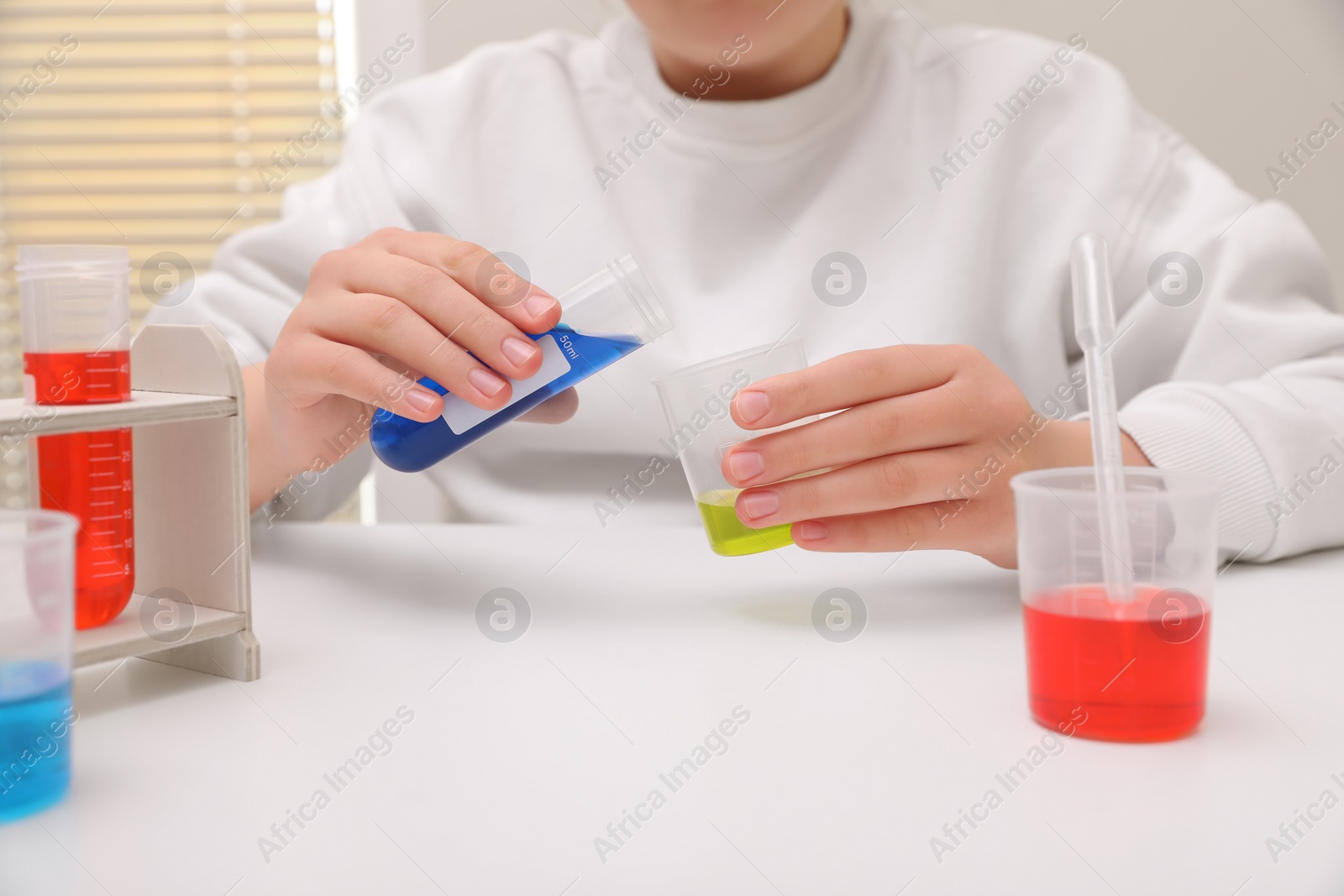 Photo of Girl mixing colorful liquids at white table indoors, closeup. Chemical experiment set for kids