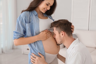 Photo of Young husband kissing his pregnant wife's tummy at home