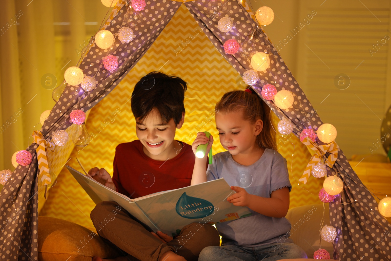 Photo of Little children with flashlight reading book in play tent at home