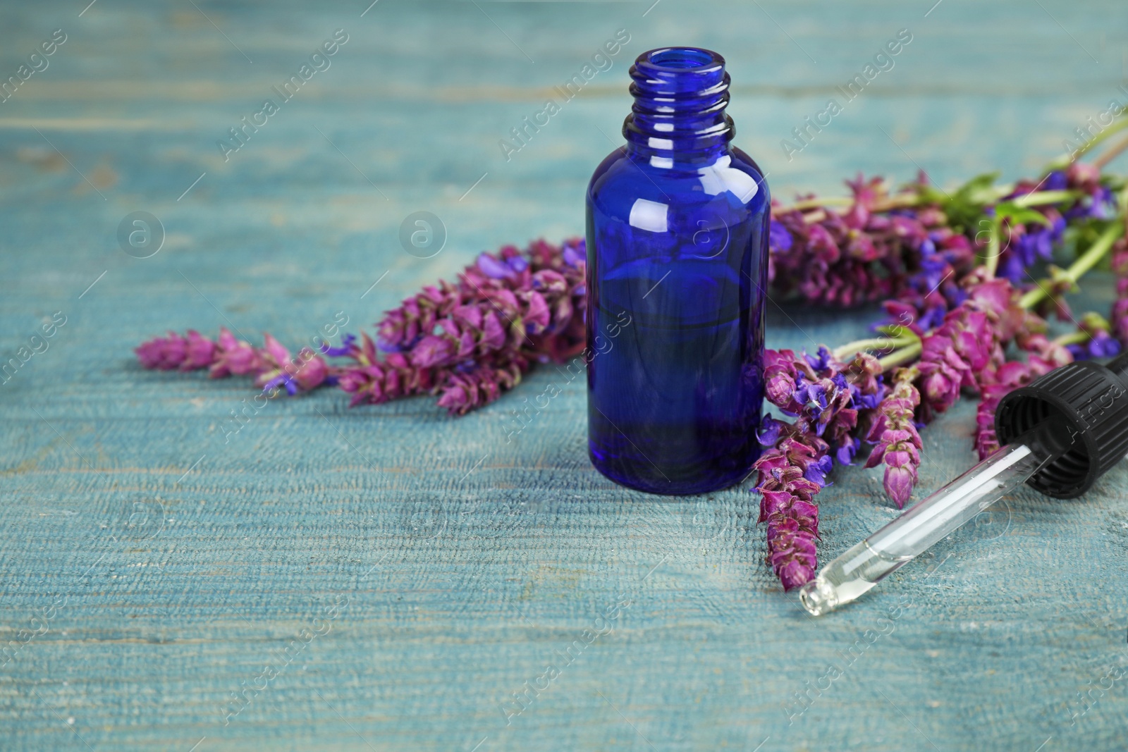 Photo of Bottle of sage essential oil and flowers on color table, space for text