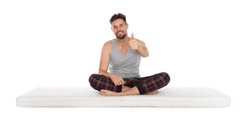 Smiling man sitting on soft mattress and showing thumb up against white background