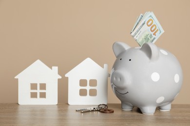 Photo of Piggy bank, keys, dollar banknotes and house models on wooden table against beige background