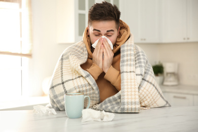 Photo of Sick young man with cup of hot drink and tissues in kitchen. Influenza virus