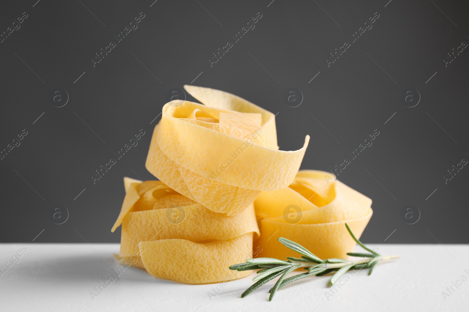 Photo of Uncooked pappardelle pasta  with rosemary on white table against dark grey background