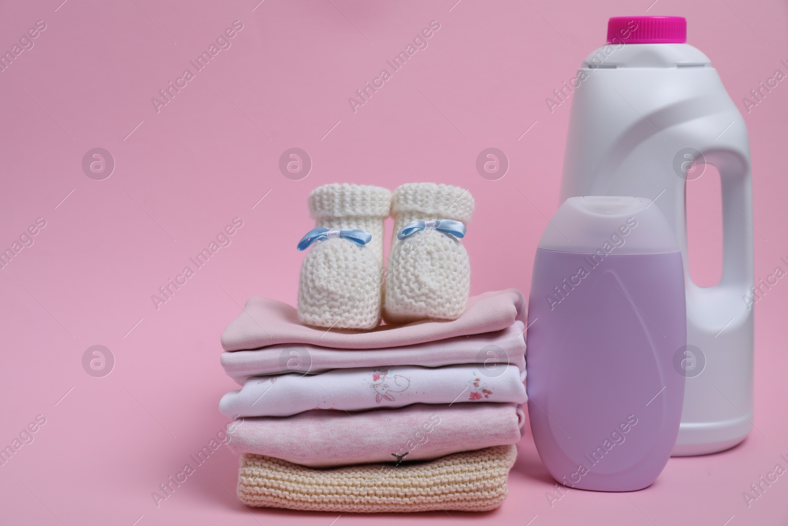 Photo of Laundry detergents, stack of baby clothes and small booties on pink background. Space for text