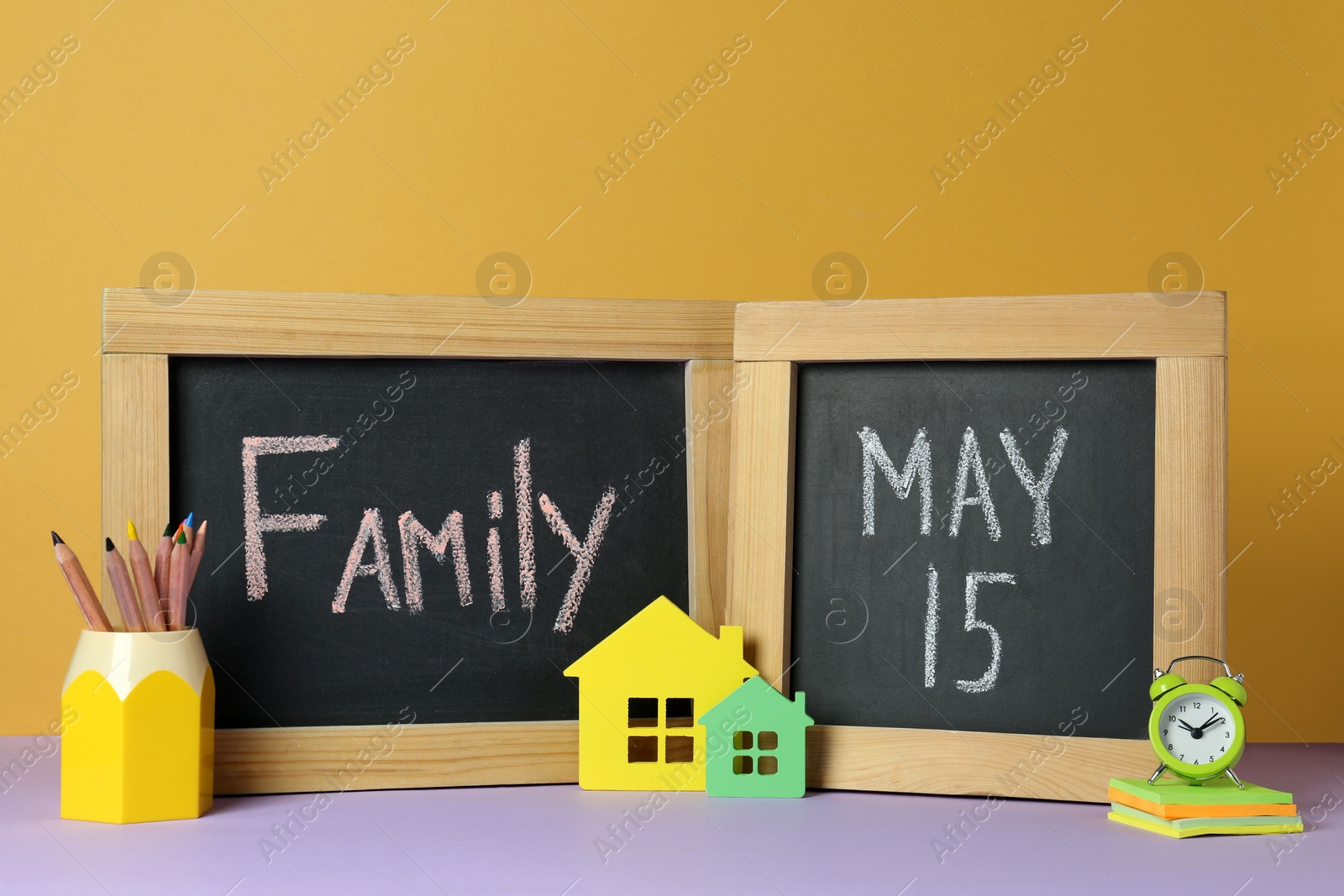 Photo of Happy International Family Day. Small chalkboards with text, house models, alarm clock and stationery on violet table against yellow background