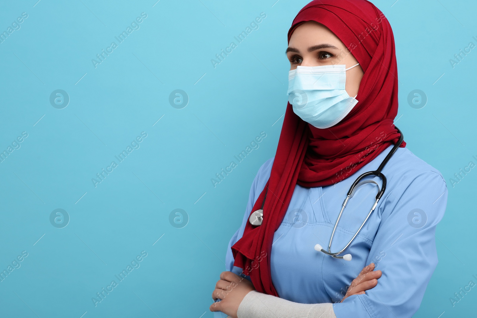 Photo of Muslim woman wearing hijab, medical uniform and protective mask on light blue background, space for text