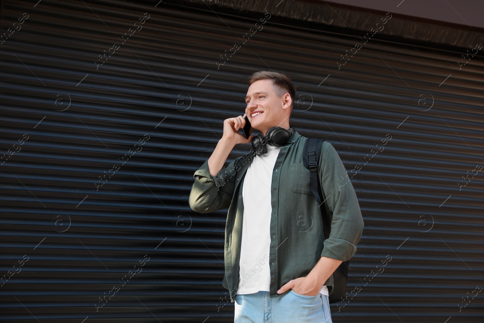 Photo of Smiling man with headphones talking on smartphone near shutters outdoors