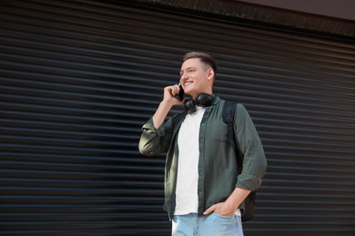 Photo of Smiling man with headphones talking on smartphone near shutters outdoors