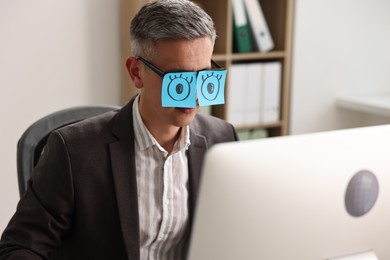 Man with fake eyes painted on sticky notes snoozing at workplace in office