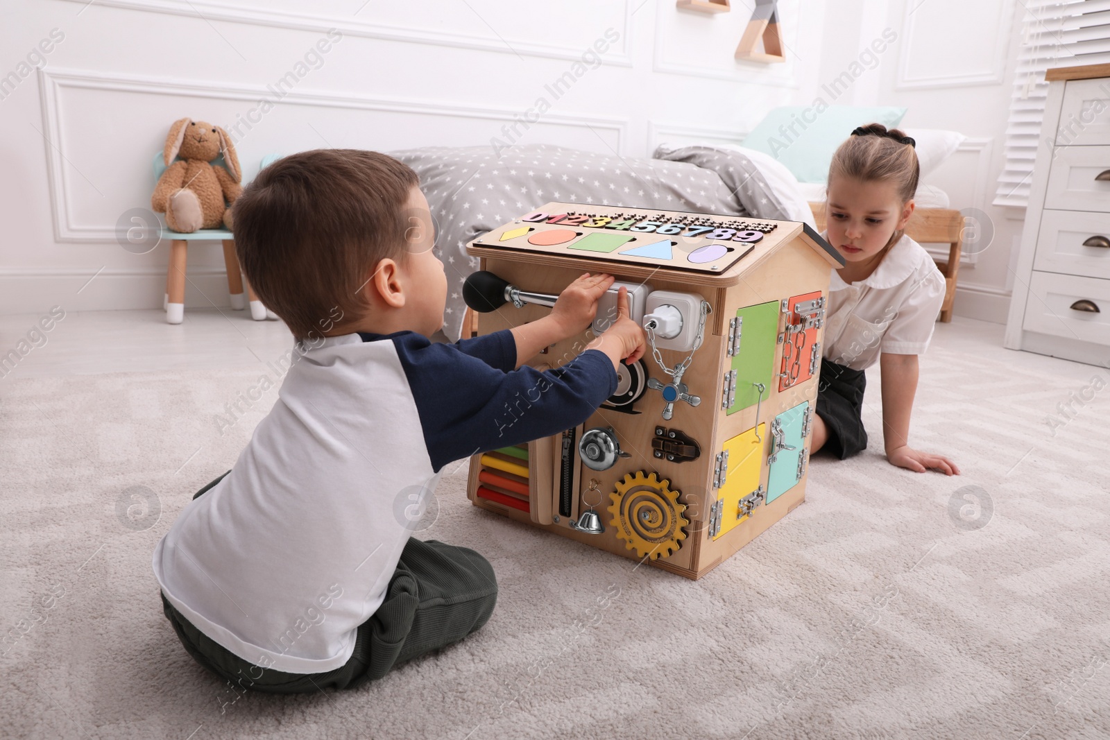 Photo of Little boy and girl playing with busy board house on floor in room