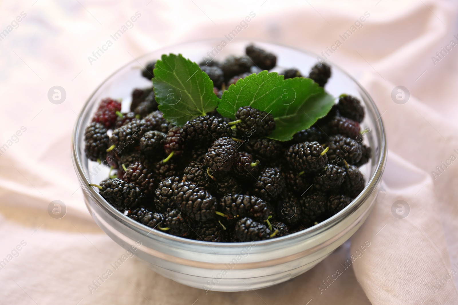 Photo of Bowl of delicious ripe black mulberries with green leaves on white fabric