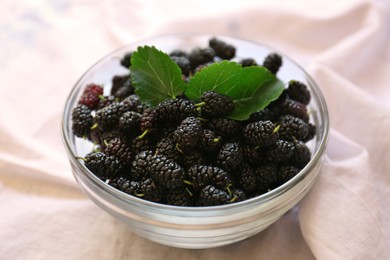 Bowl of delicious ripe black mulberries with green leaves on white fabric