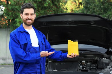 Photo of Smiling worker showing yellow container of motor oil near car outdoors