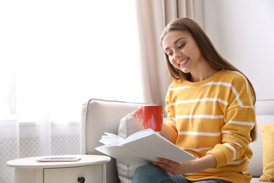 Photo of Young woman with cup of coffee reading book on couch at home