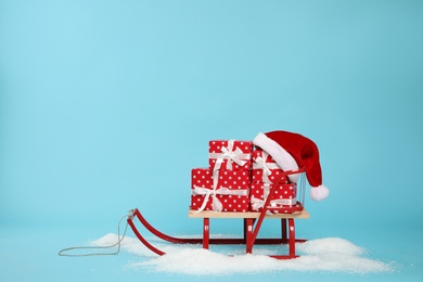 Photo of Stylish sleigh with Christmas gifts and Santa hat in pile of snow on light blue background, space for text