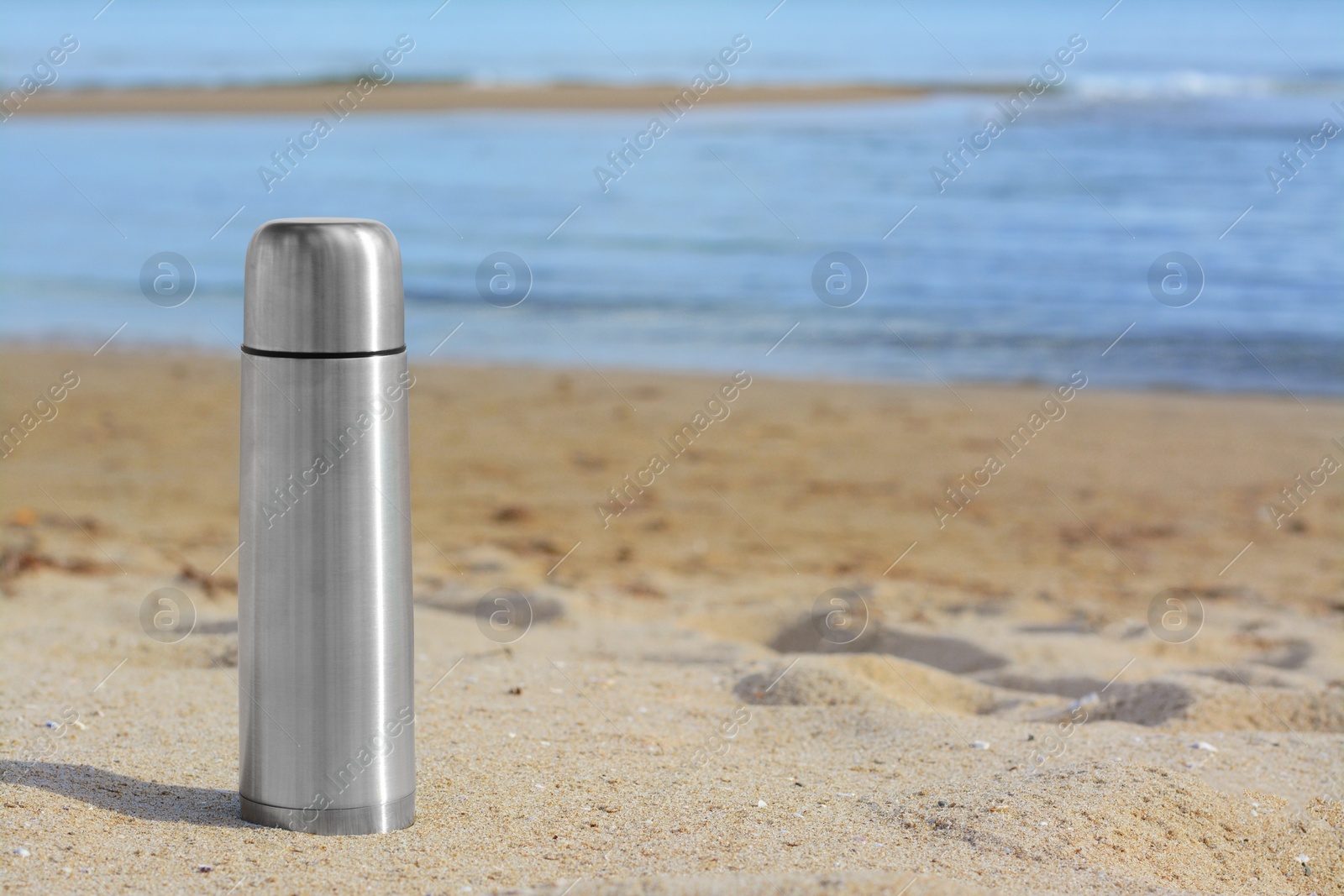 Photo of Metallic thermos with hot drink on sandy beach near sea, space for text