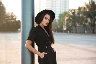 Beautiful young woman in stylish black dress and hat on city street