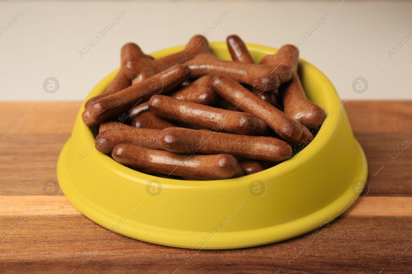 Photo of Yellow bowl with bone shaped dog cookies on wooden board, closeup