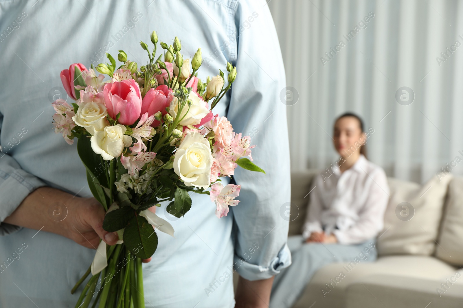 Photo of Man hiding bouquet of flowers for his beloved woman indoors, closeup