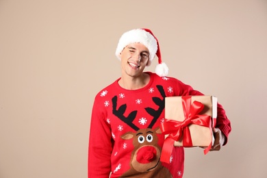 Photo of Happy man in Christmas sweater and Santa hat holding gift box on beige background