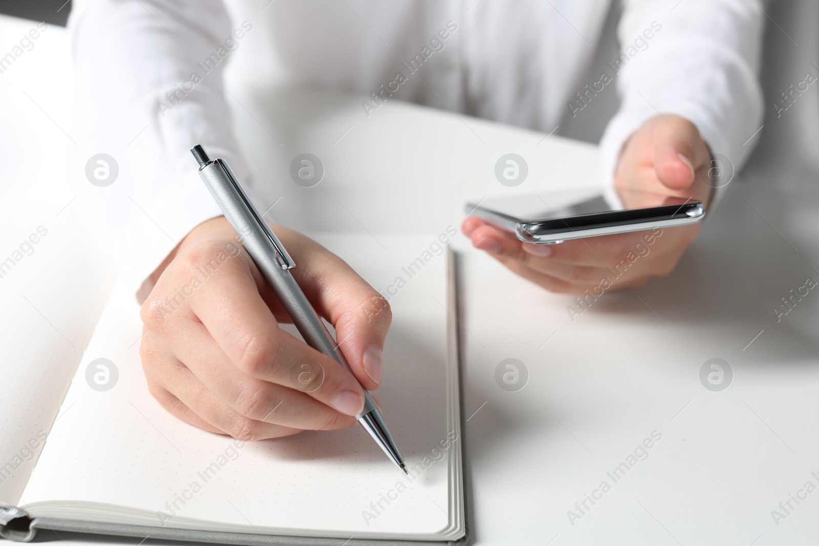 Photo of Woman with smartphone writing in notebook at white table in office, closeup