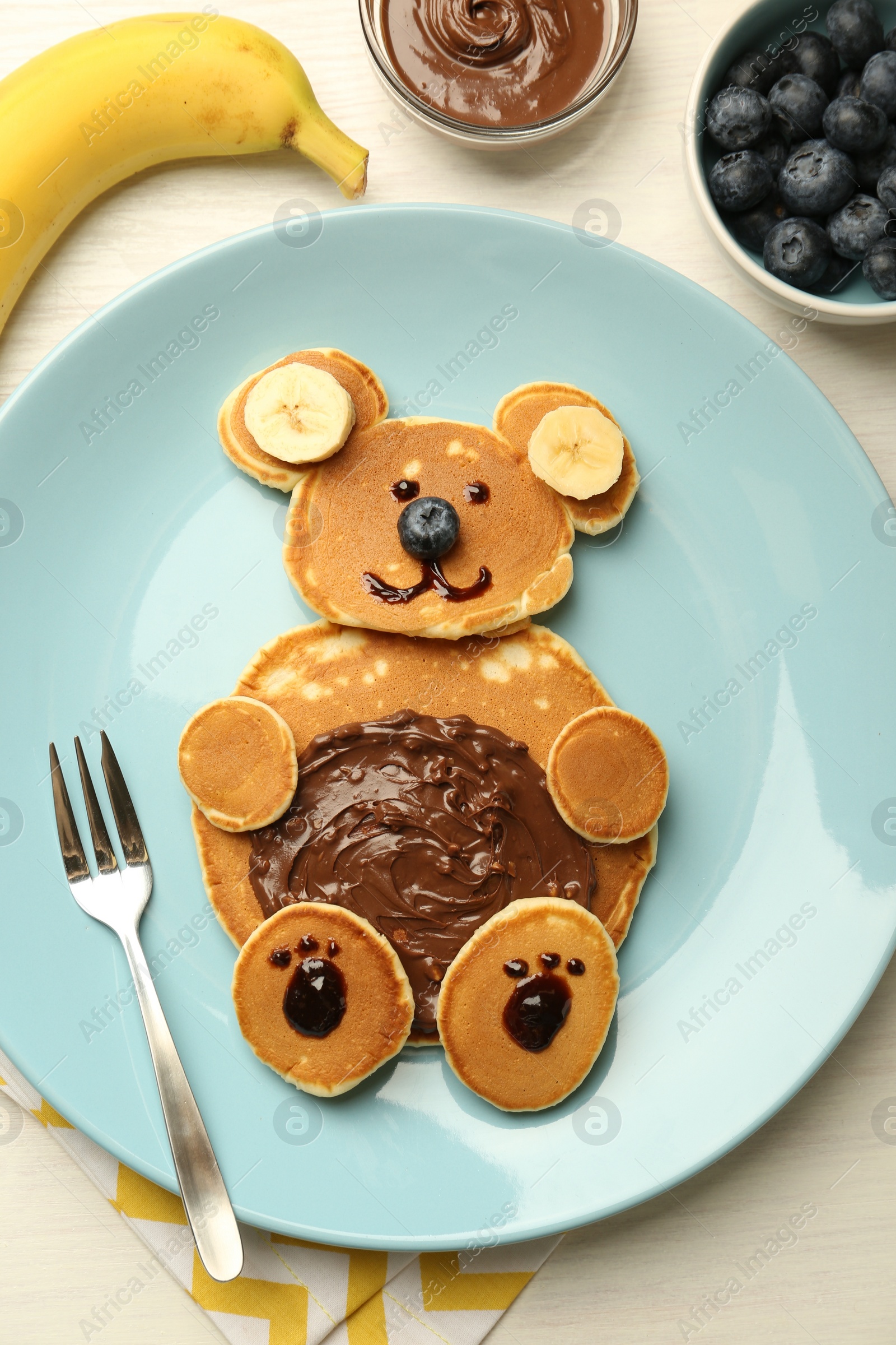 Photo of Creative serving for kids. Plate with cute bear made of pancakes, blueberries, bananas and chocolate paste on light wooden table, flat lay