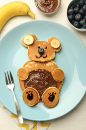 Photo of Creative serving for kids. Plate with cute bear made of pancakes, blueberries, bananas and chocolate paste on light wooden table, flat lay