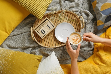 Photo of Woman holding cup of coffee while lying on bed with pillows and warm plaid, top view