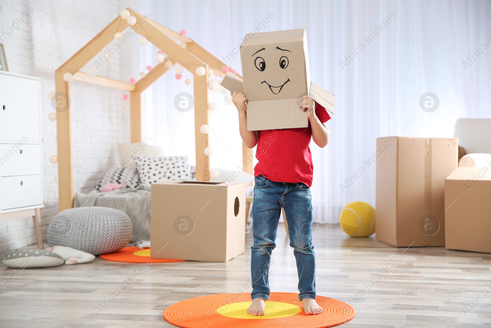 Photo of Cute little child wearing cardboard box with smiling face in bedroom