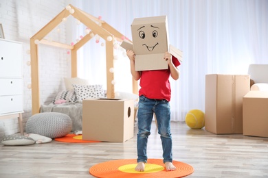Cute little child wearing cardboard box with smiling face in bedroom