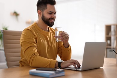 Photo of Young man with glass of water watching webinar at table in room