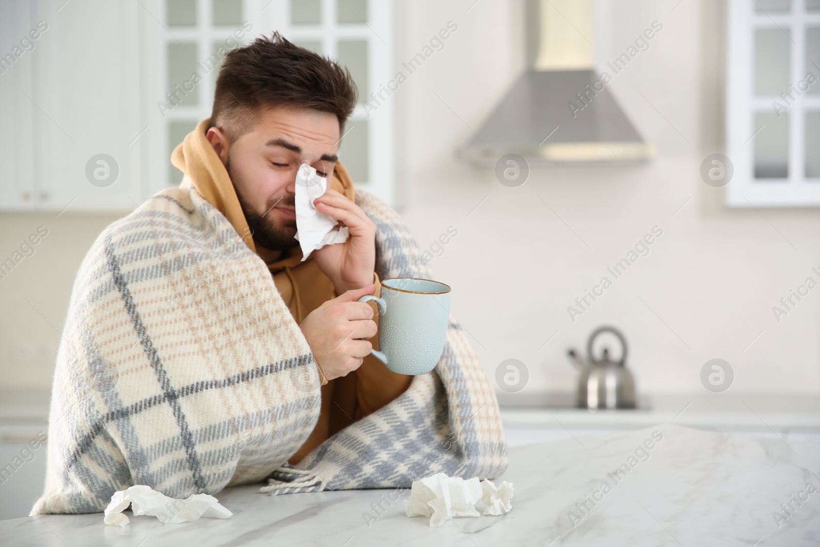 Photo of Sick young man with cup of hot drink and tissues in kitchen. Influenza virus