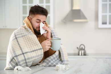 Sick young man with cup of hot drink and tissues in kitchen. Influenza virus