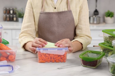 Woman sticking paper note onto container with fresh carrots at white marble table in kitchen, closeup. Food storage