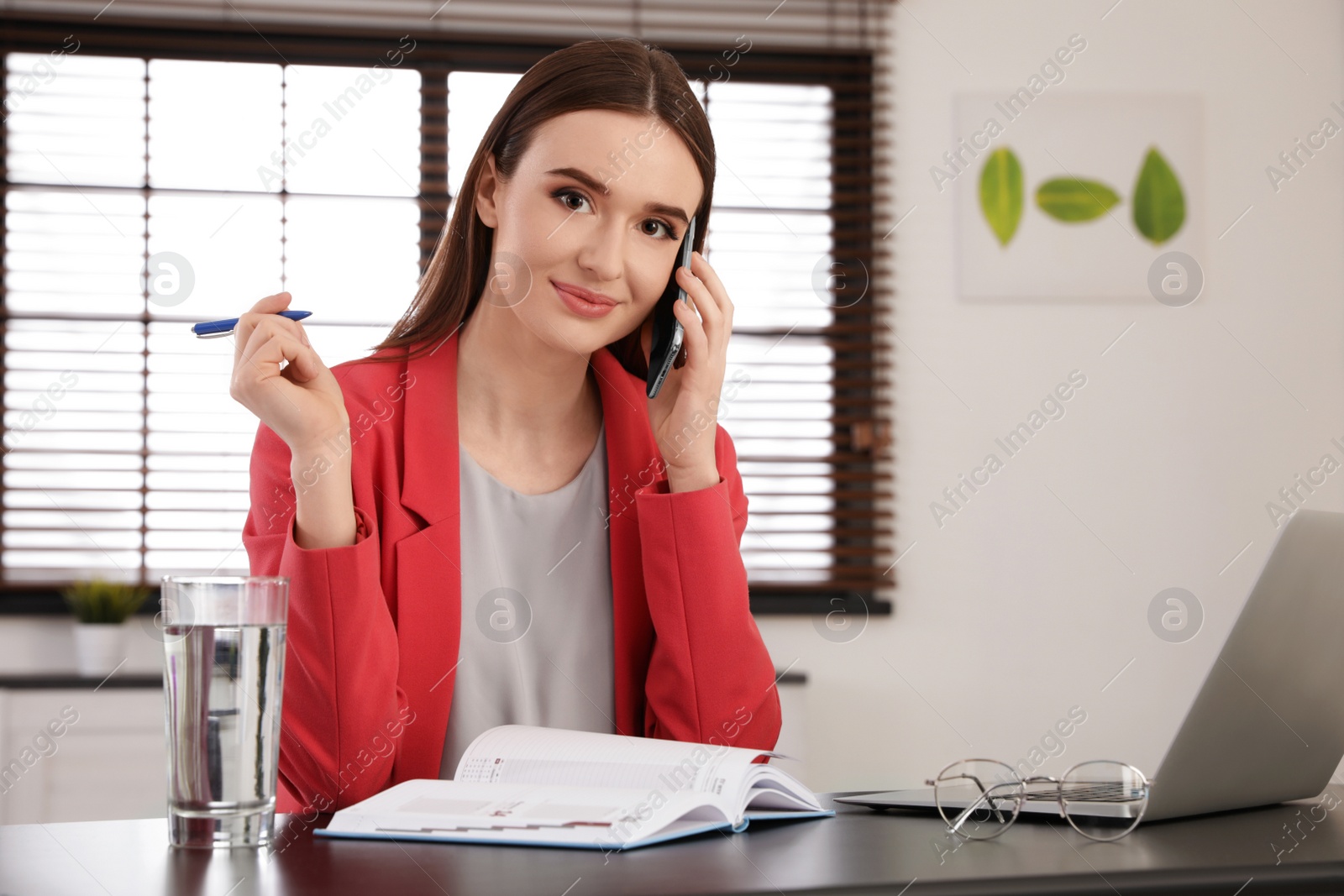 Photo of Young businesswoman talking on phone while working with notebook and laptop at table in office