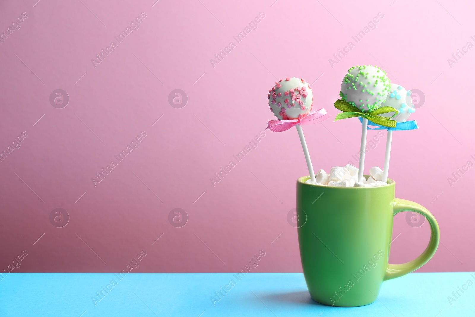 Photo of Tasty cake pops in cup with marshmallow on light blue table against lilac background. Space for text