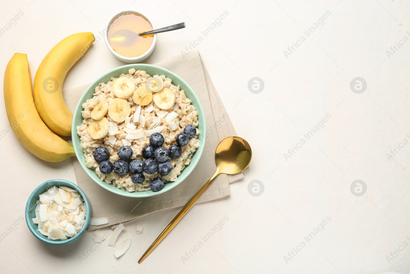 Photo of Tasty oatmeal with banana, blueberries, coconut flakes and honey served in bowl on beige table, flat lay. Space for text