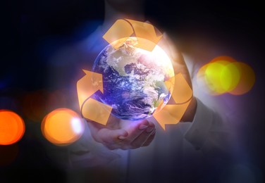Image of Woman holding virtual image of Earth with recycling symbol on dark background, closeup view. Bokeh effect