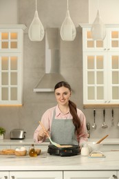 Photo of Happy woman cooking delicious crepe on electric maker at white table in kitchen