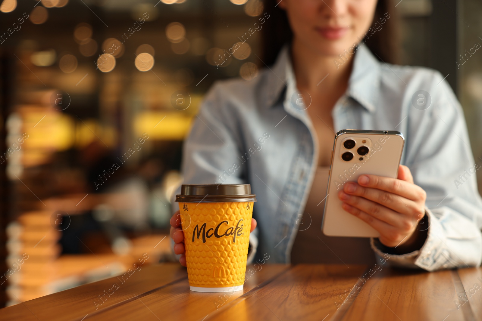 Photo of Lviv, Ukraine - September 26, 2023: Woman with hot McDonald's drink and smartphone at wooden table in cafe, closeup