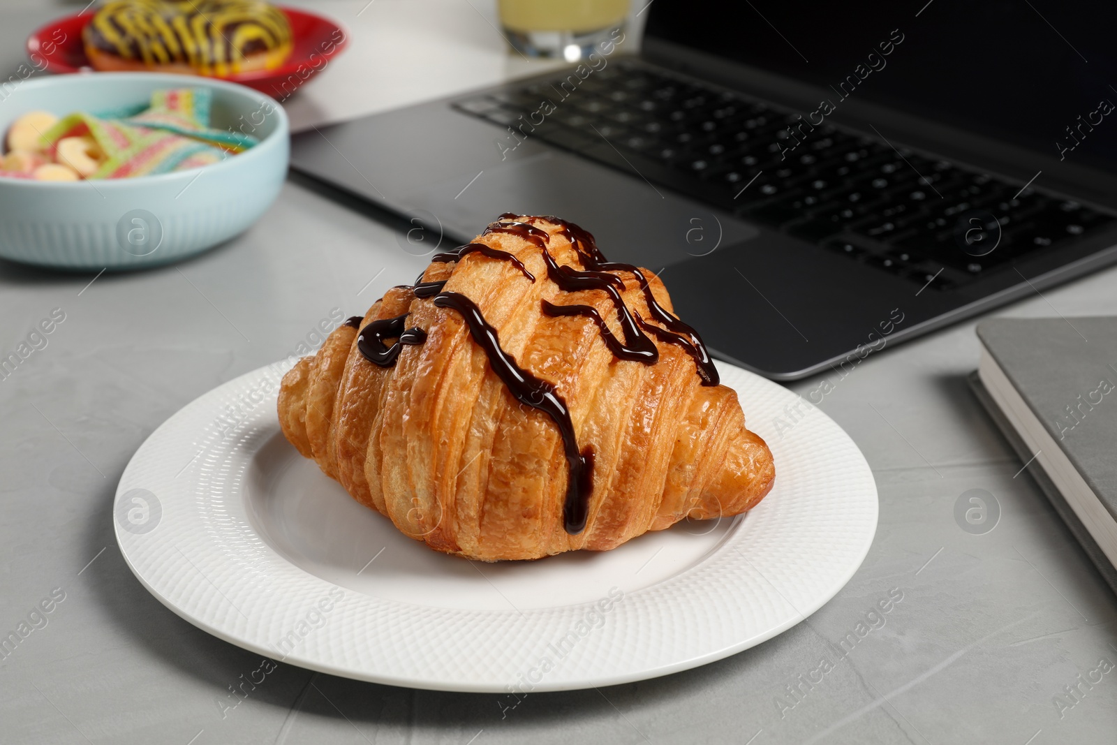 Photo of Bad eating habits at workplace. Delicious croissant and laptop on grey table, closeup
