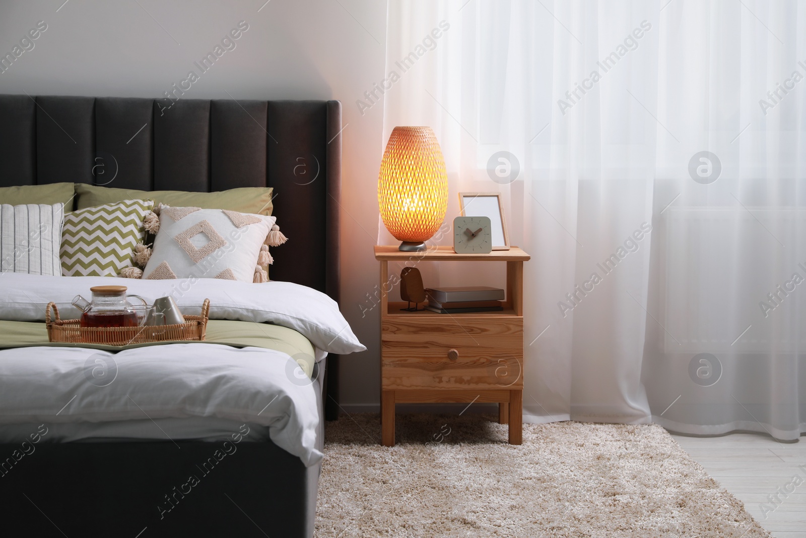 Photo of Wicker tray with teapot on comfortable bed, lamp and different decor on wooden bedside table in room. Stylish interior