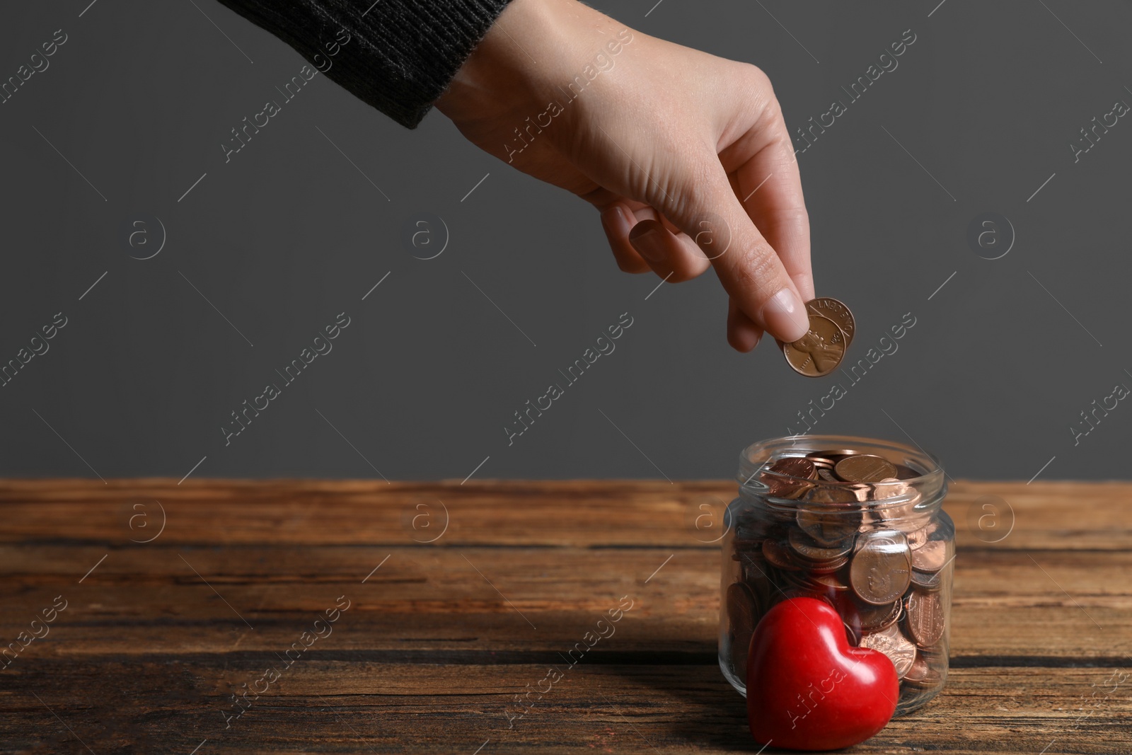 Photo of Woman putting coins into donation jar on wooden table against grey background, closeup. Space for text