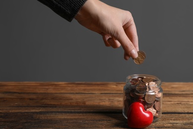 Woman putting coins into donation jar on wooden table against grey background, closeup. Space for text