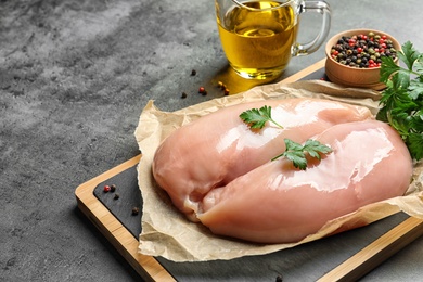 Photo of Wooden board with raw chicken breasts, pepper and parsley near olive oil in pitcher on table